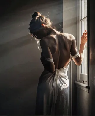 A woman stands facing away towards a window in a dimly lit room, wearing a white off-shoulder dress and her hair tied up in a bun, bathed in soft natural light.
