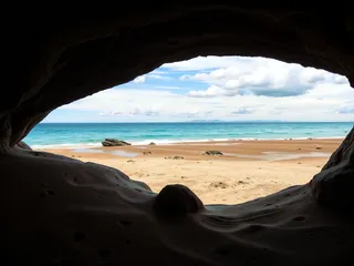View of a sandy beach and blue ocean seen through the dark opening of a rocky cave, with scattered rocks on the sand and a partly cloudy sky.