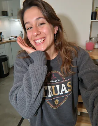 Young woman smiling brightly while wearing a gray sweater and taking a selfie indoors in a kitchen setting.