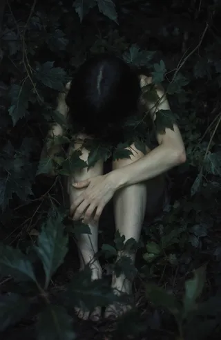 A person with light skin and short dark hair sits nude, knees drawn up, partially obscured by dense dark green foliage in dim, moody lighting.