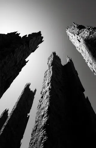Black and white photograph of abstract towering structures with jagged, textured edges viewed from a low angle, highlighting dramatic shadows and light contrasts.