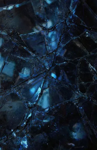 Close-up of a cracked reflective glass floor illuminated by glowing blue light from above, creating a vibrant starburst pattern across dark blue and black shattered surfaces.