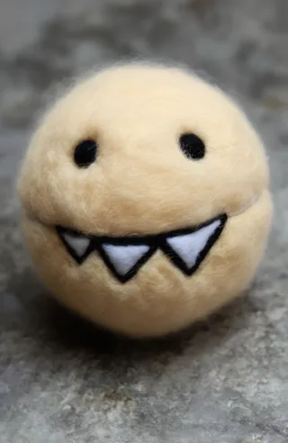 Close-up of a small round beige fuzzy ball made of felt with black circular eyes and a wide toothy grin featuring three white triangular teeth outlined in black on a blurred gray surface.