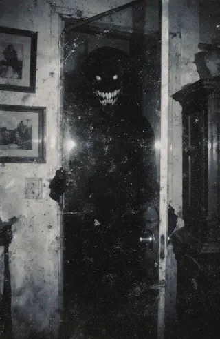 Black and white photograph of a dimly lit room with a shadowy figure featuring a wide toothy grin standing behind a scratched and smudged glass door, gripping the handle.