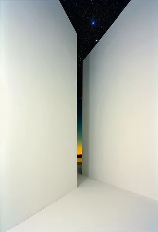 Narrow corridor formed by large white walls opening to a view of a colorful sunset gradient and a star-filled night sky above.