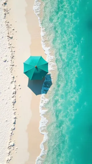Aerial view of a tranquil beach showing a single turquoise umbrella casting a shadow on soft white sand near the clear blue water with gentle waves.
