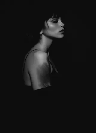 Black and white side profile portrait of a woman with short hair, earrings, and visible tattoos on her arm, set against a dark background.