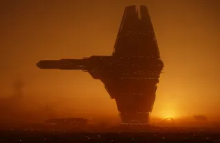 Silhouetted towering spaceship against an orange sunset sky in a sci-fi desert landscape with glowing lights and smaller ships in the background.