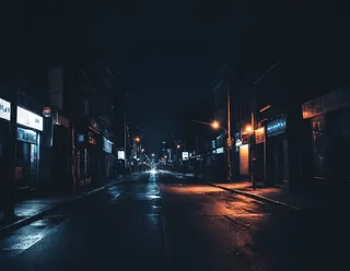 A dark urban city street at night illuminated by street lights reflecting on wet pavement with a deep dark background.