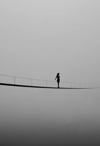 Black silhouette of a figure standing on a narrow bridge or tightrope suspended over thick white mist, with minimal details and a spacious foggy background.