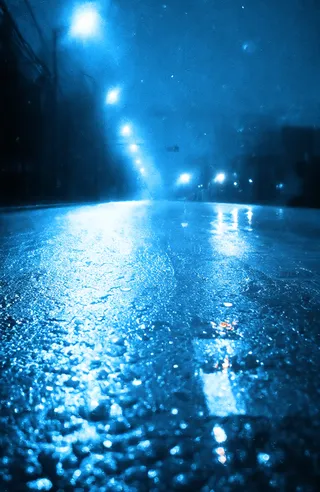 Wet urban street at night illuminated by bright blue streetlights on a rainy night with reflections on the wet road and a misty, moody atmosphere.