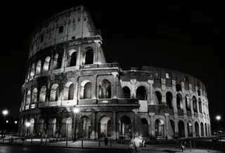 Black and white night photograph of a massive ancient colosseum, half-ruined with arches and columns, illuminated with dramatic lighting and fog under a starry sky.