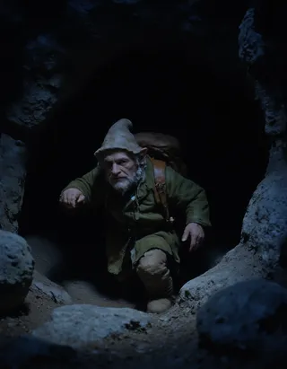 A gnome with a white beard and pointy hat wearing green clothes and a brown leather backpack climbing out of a dark cave at night.