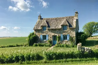 Stone cottage with blue shutters surrounded by green fields and flowering garden under a clear blue sky in the French countryside