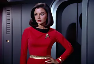 Young woman with shiny voluminous long bob haircut dressed as a redshirt yeoman security in Star Trek Original Series style, standing in Starship Enterprise corridor.