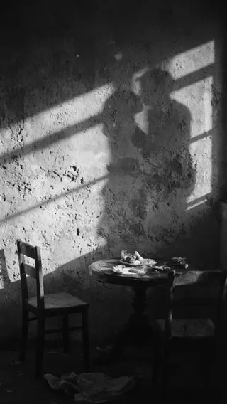 Black and white photograph showing a shadow silhouette of a couple kissing cast on a peeling plaster wall, with a rustic round table and two wooden chairs in a sunlit room
