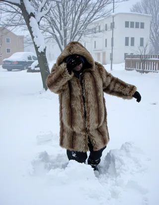 Man wearing a large fur coat with hood covering his face while walking through knee deep snow during a blizzard, side view.
