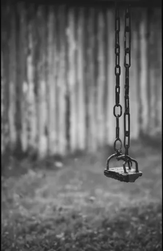 Black and white photograph of a rusty metal swing suspended by chains with raindrops, positioned against a blurred wooden fence background with soft, diffused lighting.