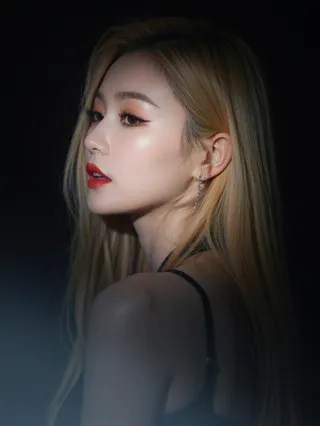Close-up side profile of a female K-pop star with long blonde hair and bold red lipstick, dramatically lit against a pitch-black background with a narrow spotlight.