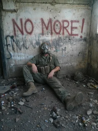 A soldier wearing a dusty gas mask and worn military uniform sits exhausted against a cracked, graffiti-covered concrete wall with 'NO MORE!' painted in red above him.