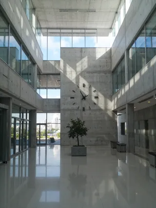 Interior lobby of a contemporary office building with Brutalist concrete walls, large minimalist wall clock, polished floor, natural light from floor-to-ceiling windows, and an indoor tree planter.