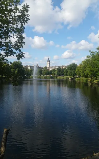 Scenic lake with a fountain in the center, framed by oak tree branches, with an American style town featuring a clock tower and buildings under a blue sky with clouds